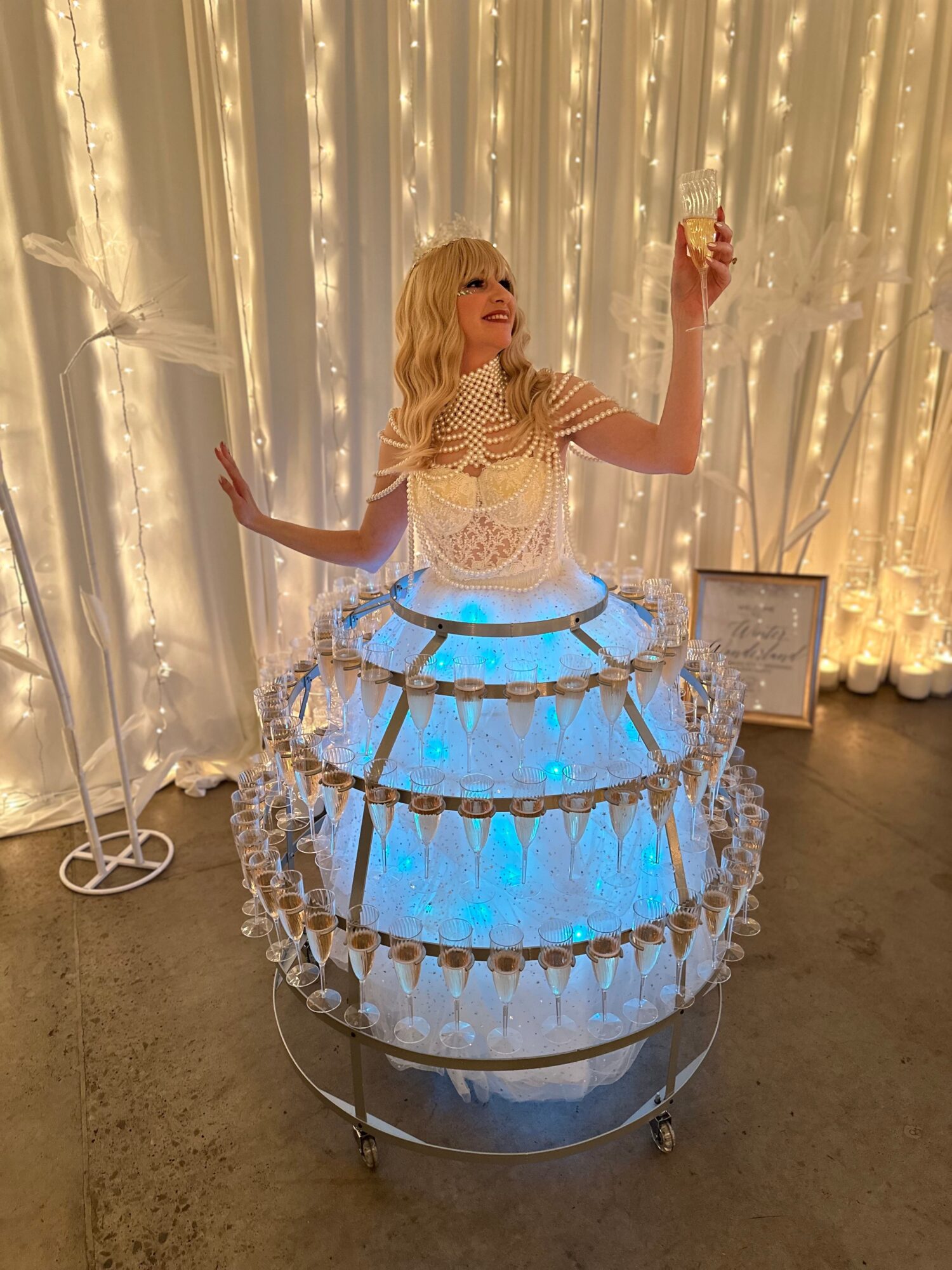 Woman in a layered, illuminated dress with a backdrop of fairy lights, holding a glass, smiling, in a decorated room.