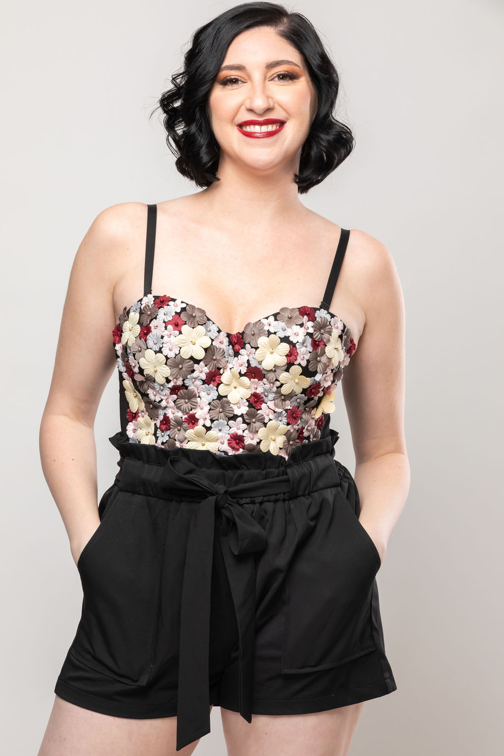 Young woman with short black hair smiling, wearing a floral top and black shorts with a bow, standing against a plain background.