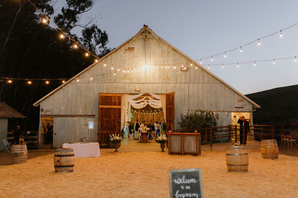 Barn decorated for an event with string lights, barrels, and a welcome sign, evening setting with people inside.