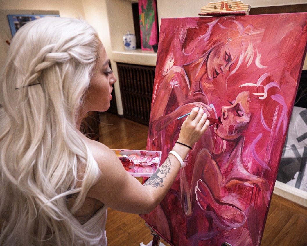 Woman with long, platinum blonde hair painting a pink and red abstract portrait on a canvas.