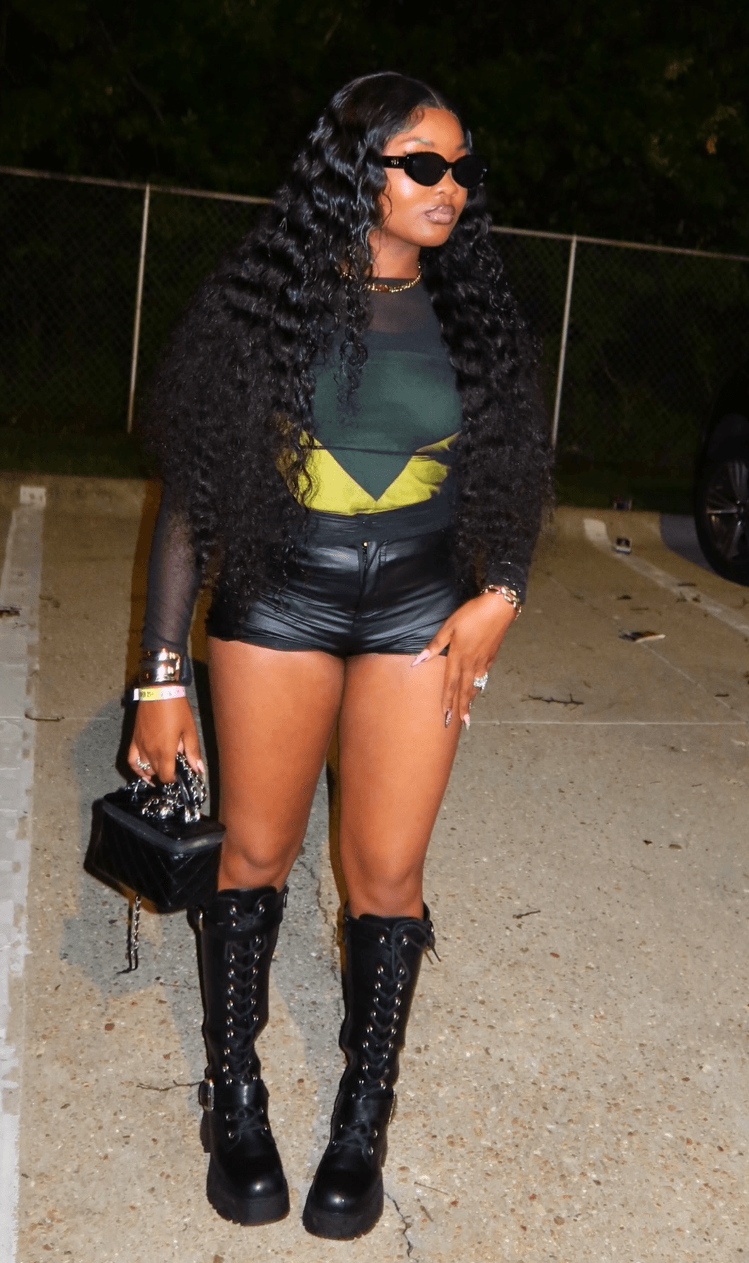 Woman with long curly hair wearing sunglasses, a black and yellow top, black shorts, and tall black boots, standing outdoors at night.