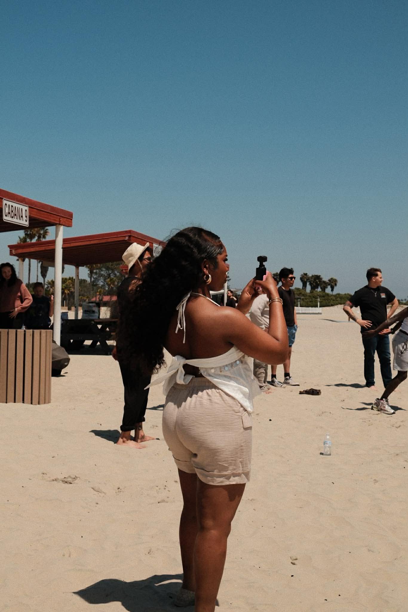 Woman with long hair taking a photo on a sandy beach, people in the background, clear blue sky.