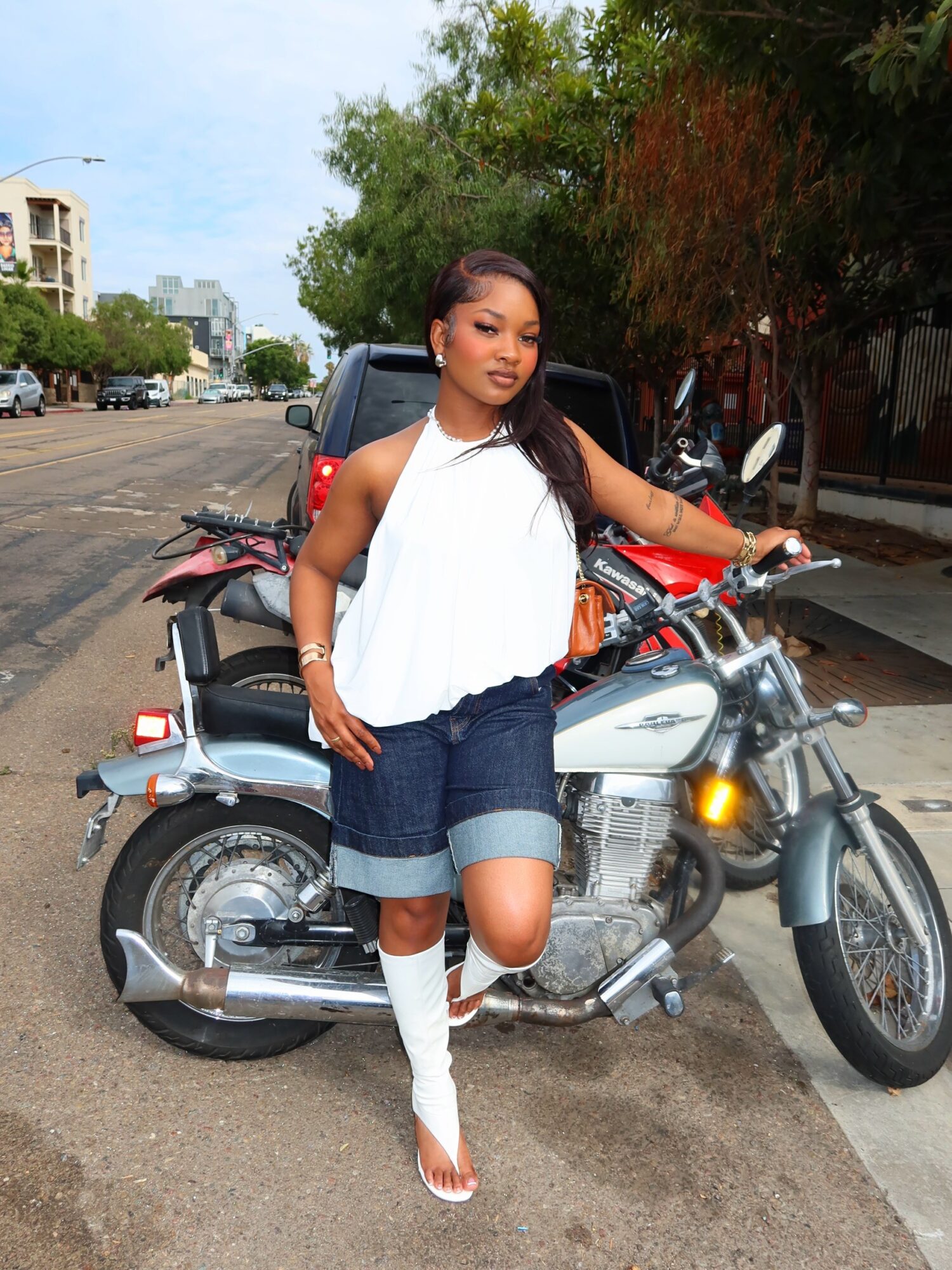 Woman standing next to a motorcycle on a city street, with trees and buildings in the background.