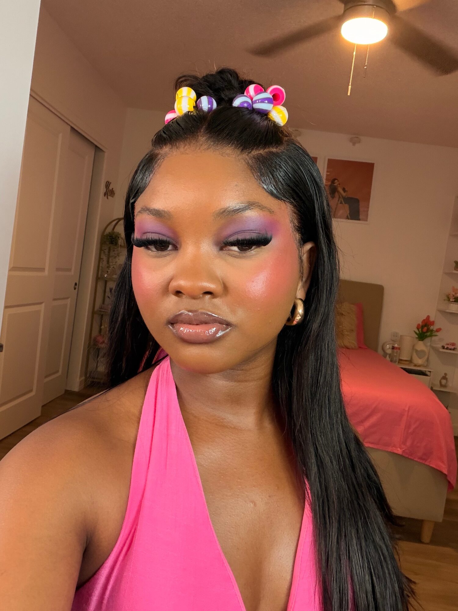 Young woman with long black hair, wearing pink top, makeup, and colorful hair accessories, in a bedroom.