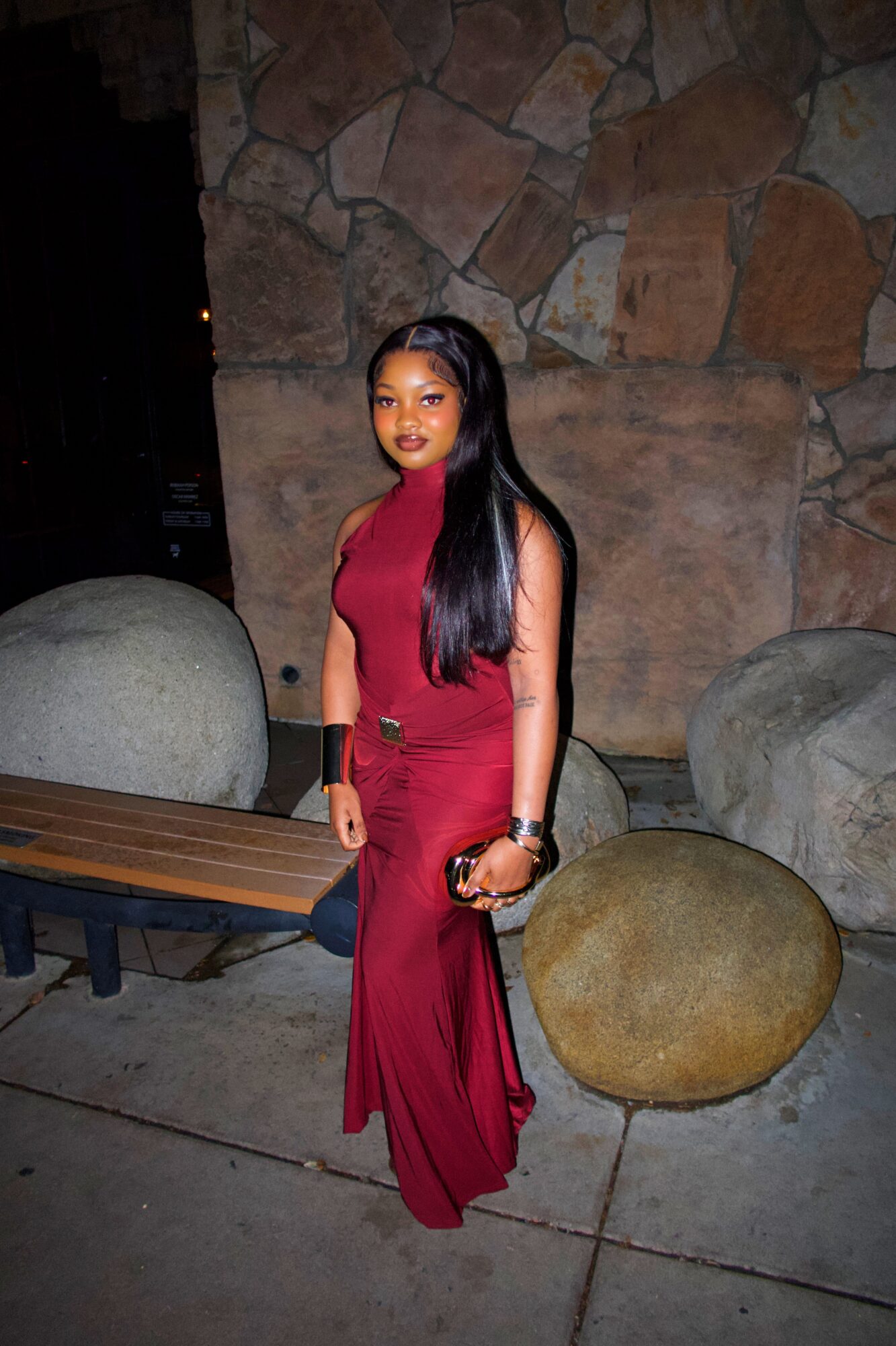 Woman in a long red dress standing outdoors near large rocks and a wooden bench at night.