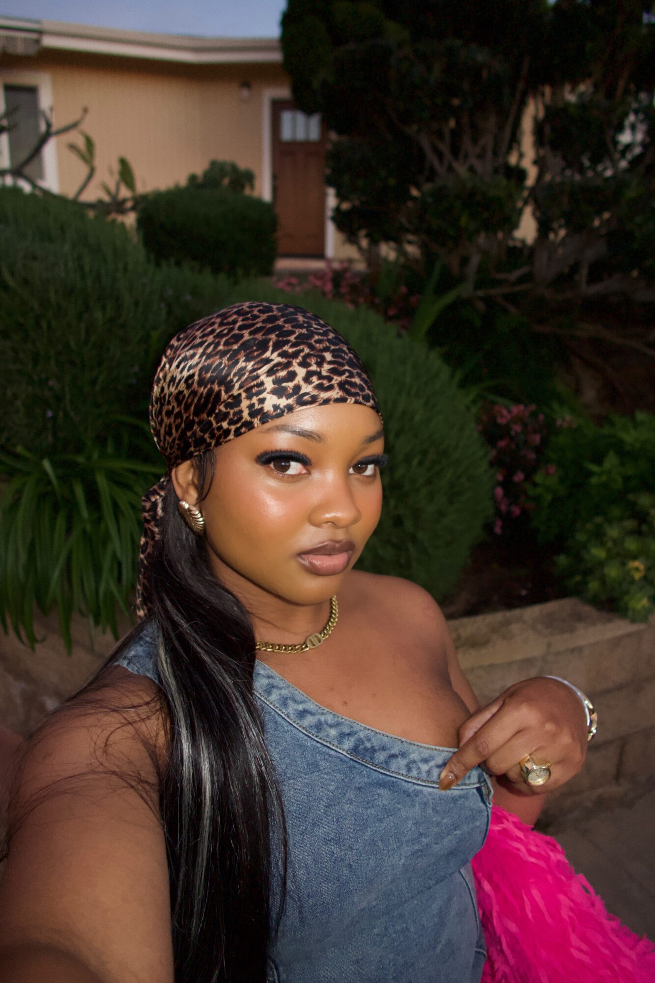 Young woman with long black hair wearing a leopard print headscarf, denim top, and pink skirt outdoors, looking at camera.