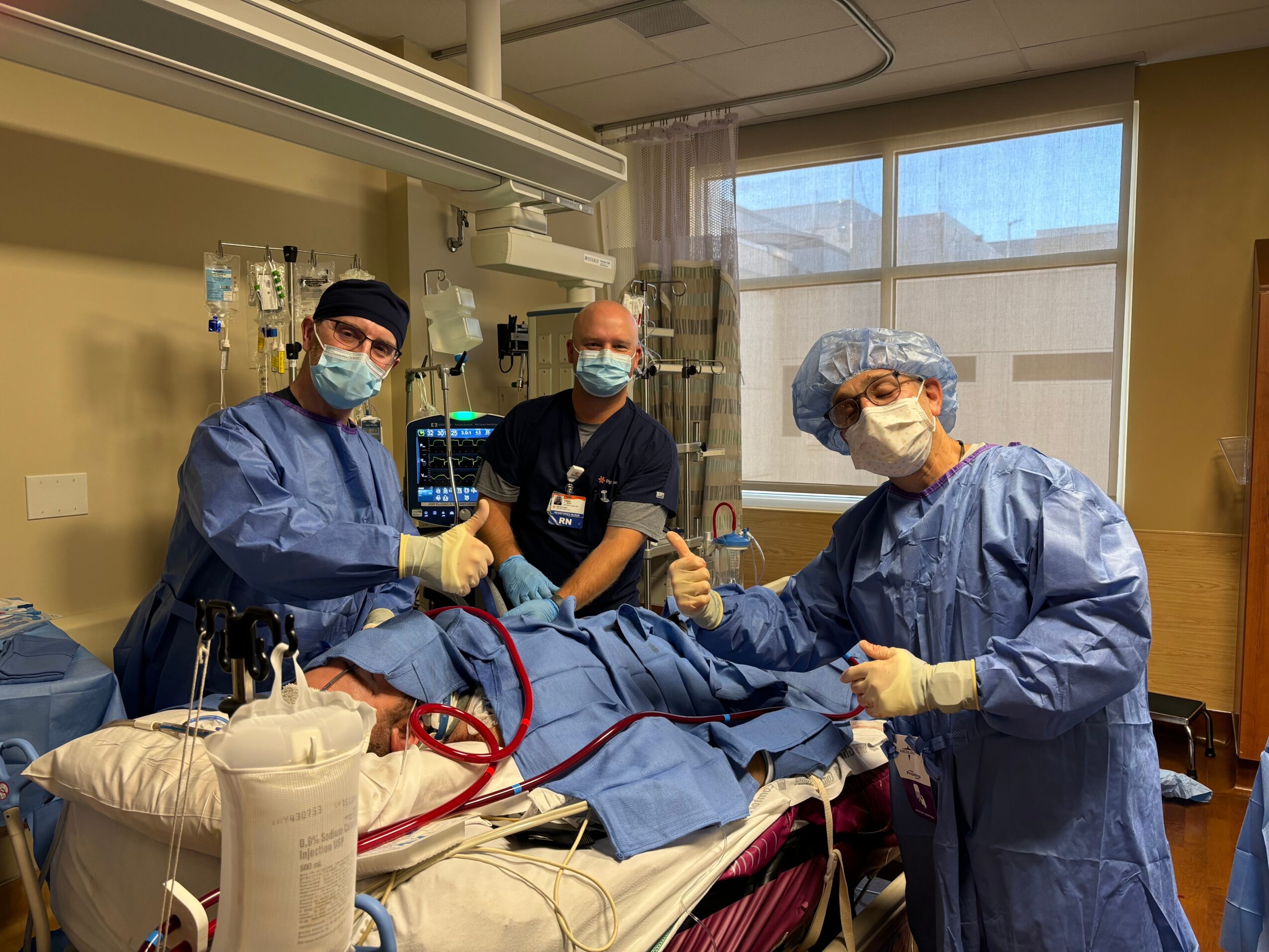 Three medical professionals in scrubs and masks give a thumbs-up around a patient on a hospital bed in a clinical room.