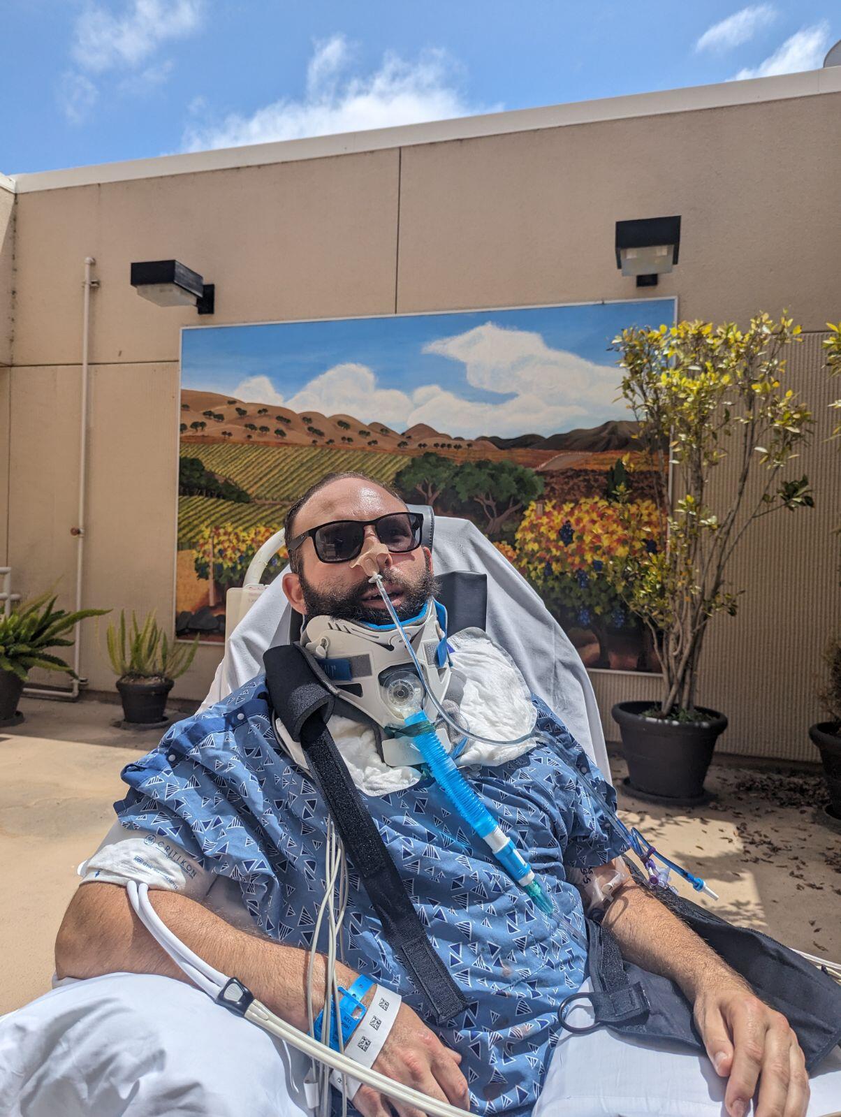 Man in hospital gown with medical equipment, sitting outdoors with a background of a wall, potted plants, and a painted landscape mural.
