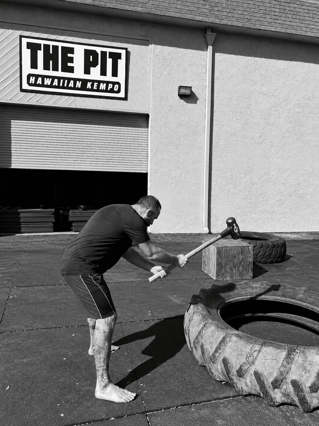 Man using sledgehammer to break a large tire outside a gym with a sign reading 'The Pit Hawaiian Kempo'.