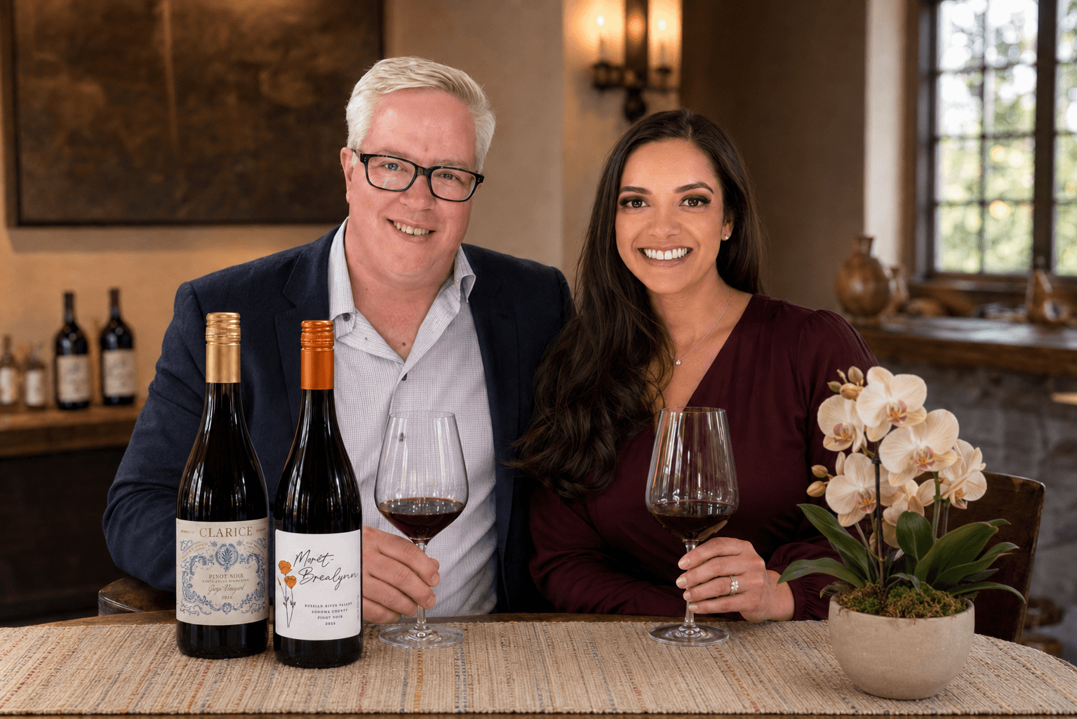 Two people sitting at a table with wine bottles and glasses, smiling, in a cozy room with a window and a plant.