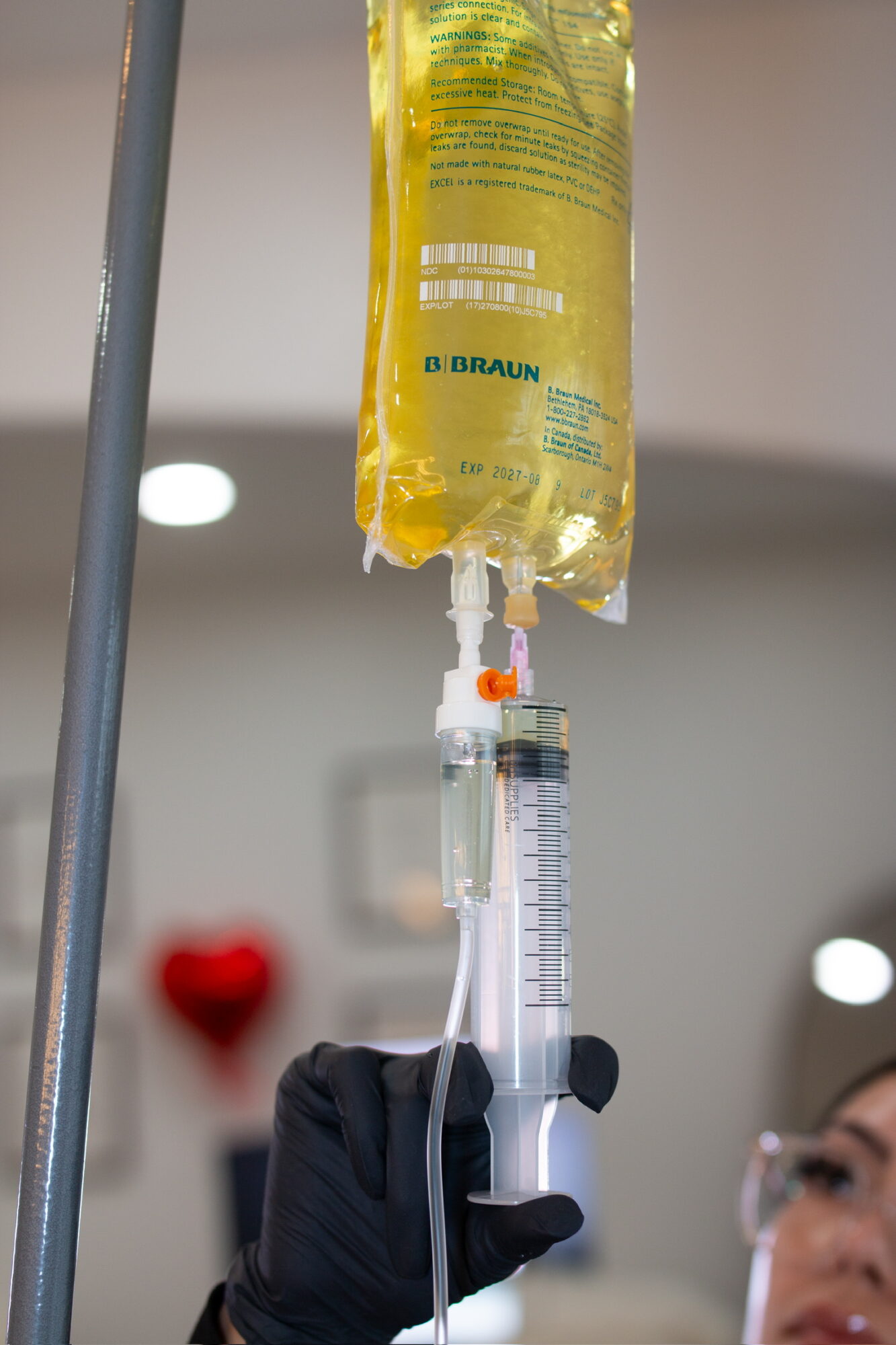 Medical IV drip bag hanging with a syringe connected, person in background, hospital setting, bright lighting.