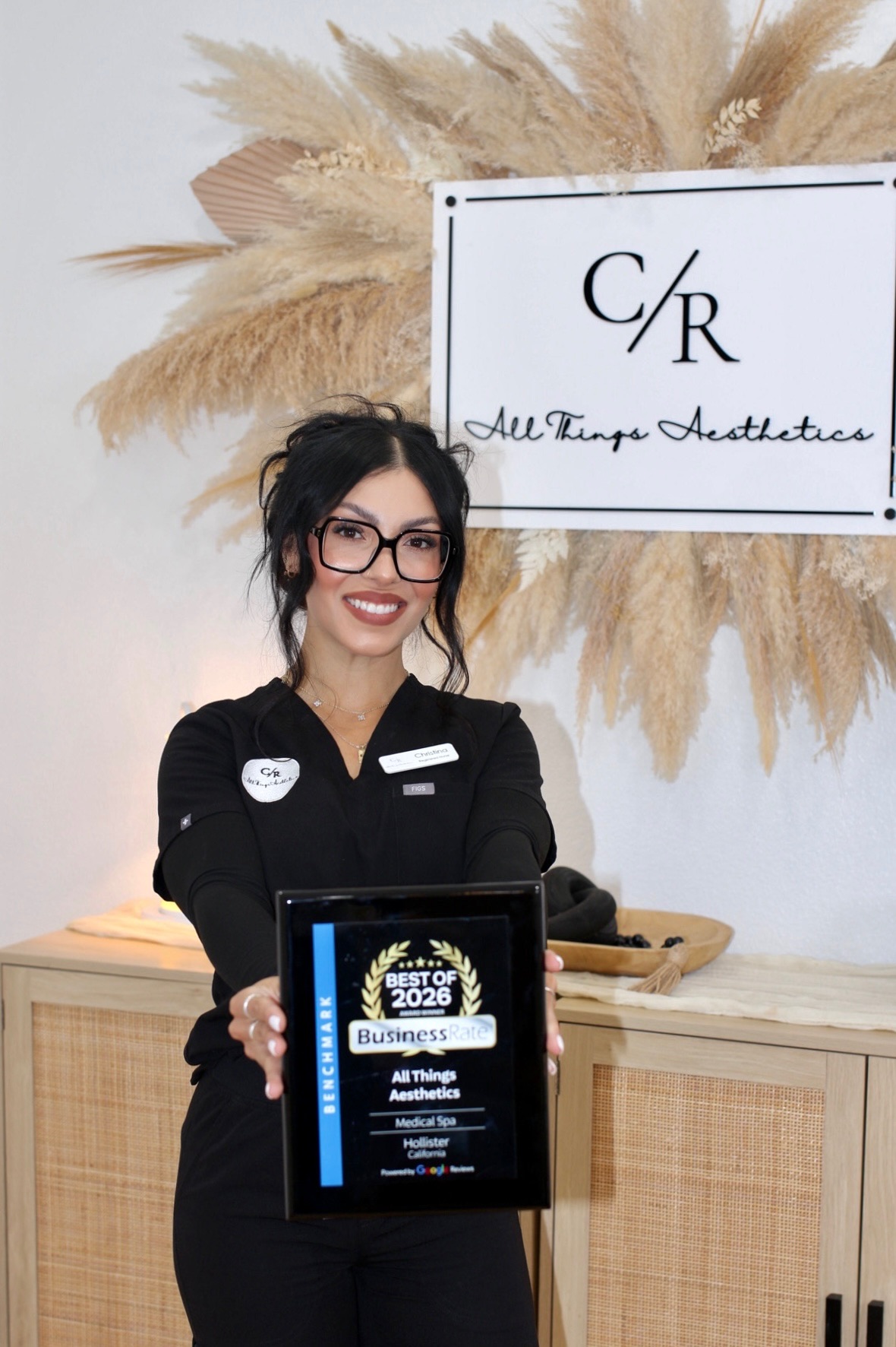 Woman with glasses and dark hair holding a framed certificate, standing in front of a sign and decorative plants.