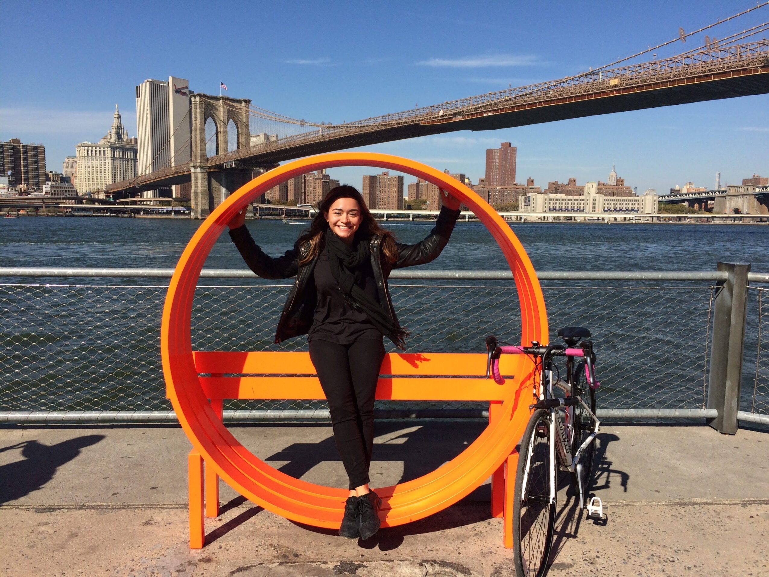 Woman standing inside a large orange circular bike rack near a river with city buildings and a bridge in the background.