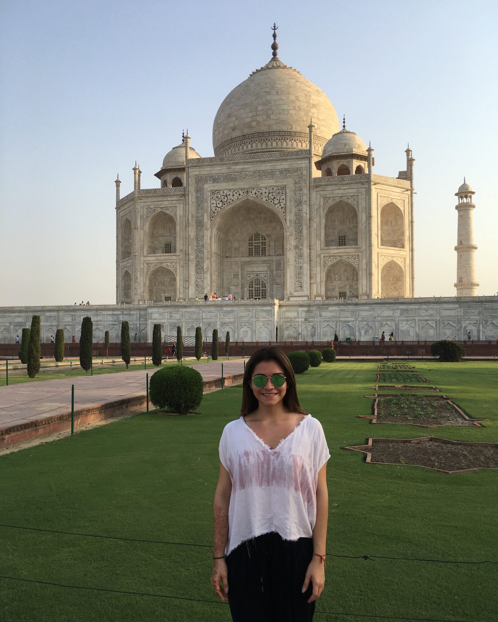 Woman with sunglasses standing in front of the Taj Mahal, green lawn and clear sky visible.