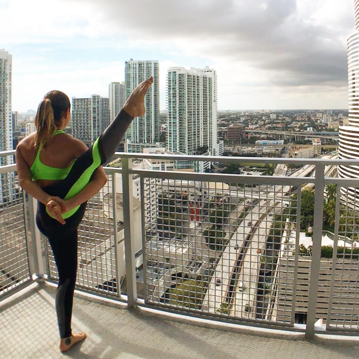 Person practicing yoga on balcony overlooking city skyline with tall buildings and cloudy sky.