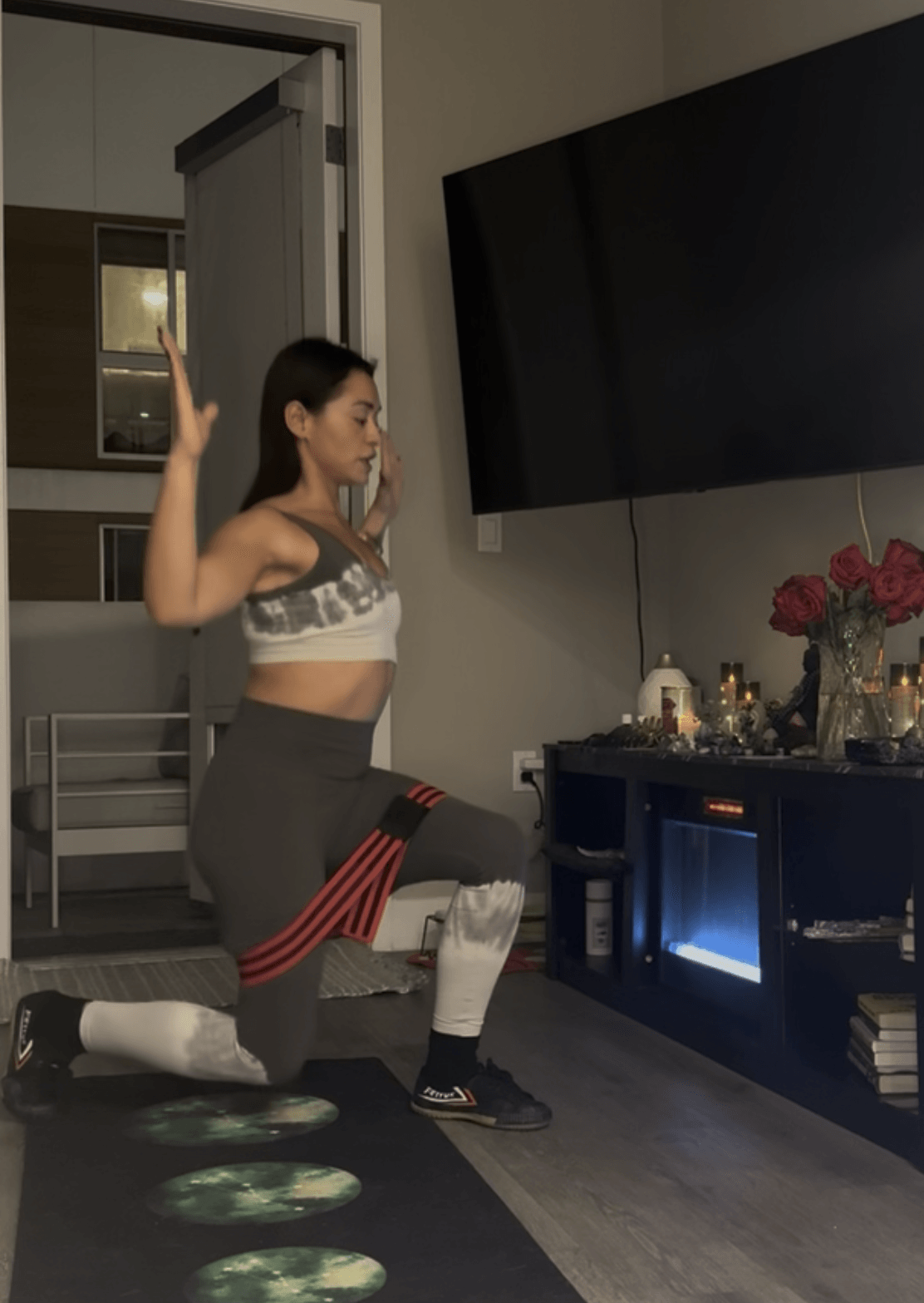 Woman in workout clothes kneeling with one arm raised in a living room, next to a TV and flowers, facing left.