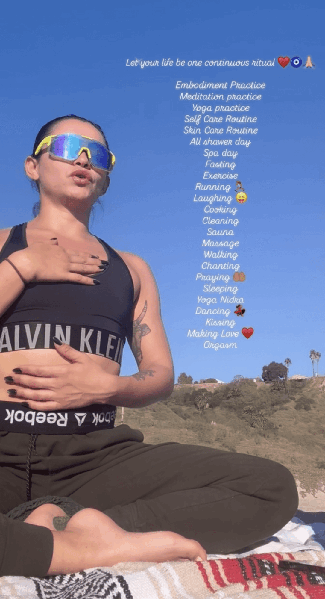 Woman sitting outdoors on a blanket, wearing sunglasses and a black top, with a blue sky background and text overlay.