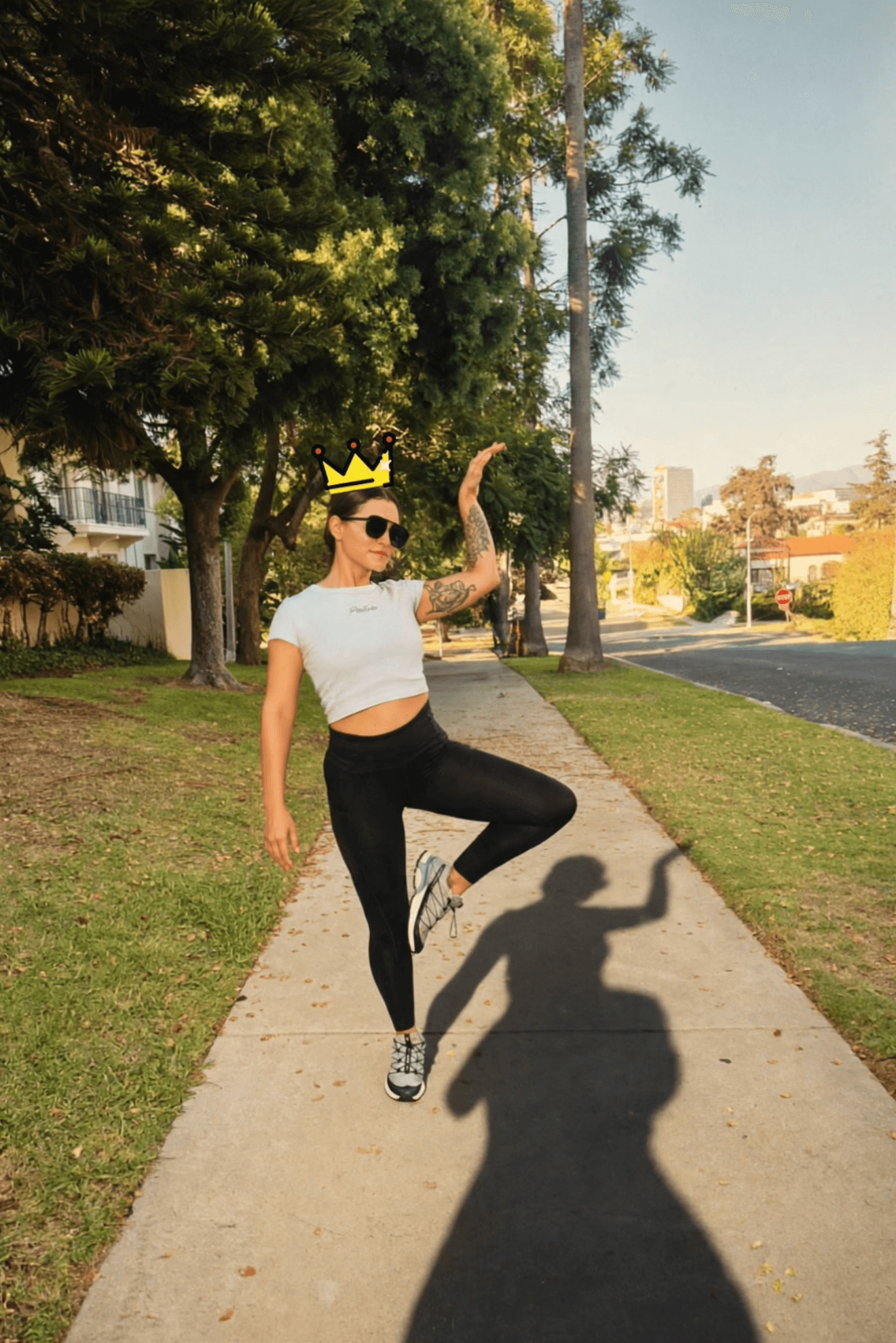 Person wearing a crown, sunglasses, white crop top, and black pants, standing on a sidewalk with one leg raised, shadow cast on ground.