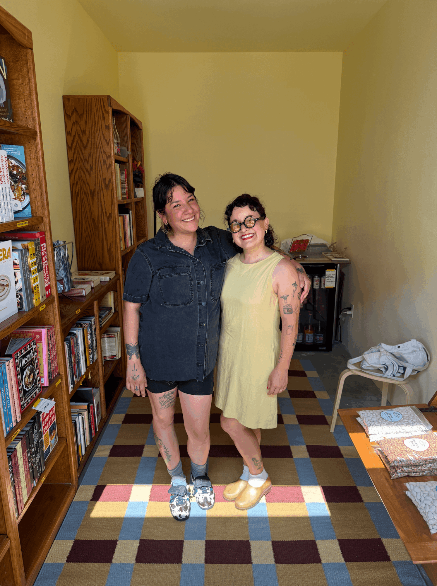 Two women stand together in a room with bookshelves, smiling, one wearing a dark shirt and shorts, the other in a yellow dress.