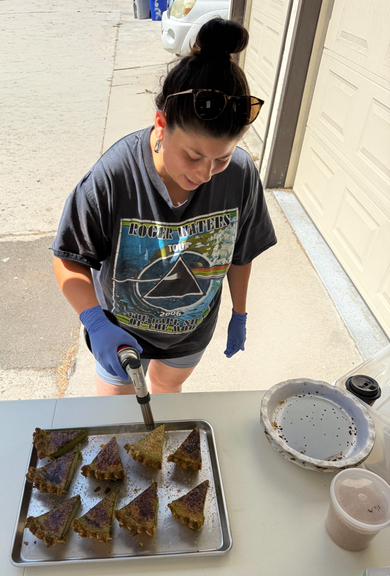 Woman wearing sunglasses and gloves, baking triangular snacks on a tray outside, with a plate and a cup nearby.