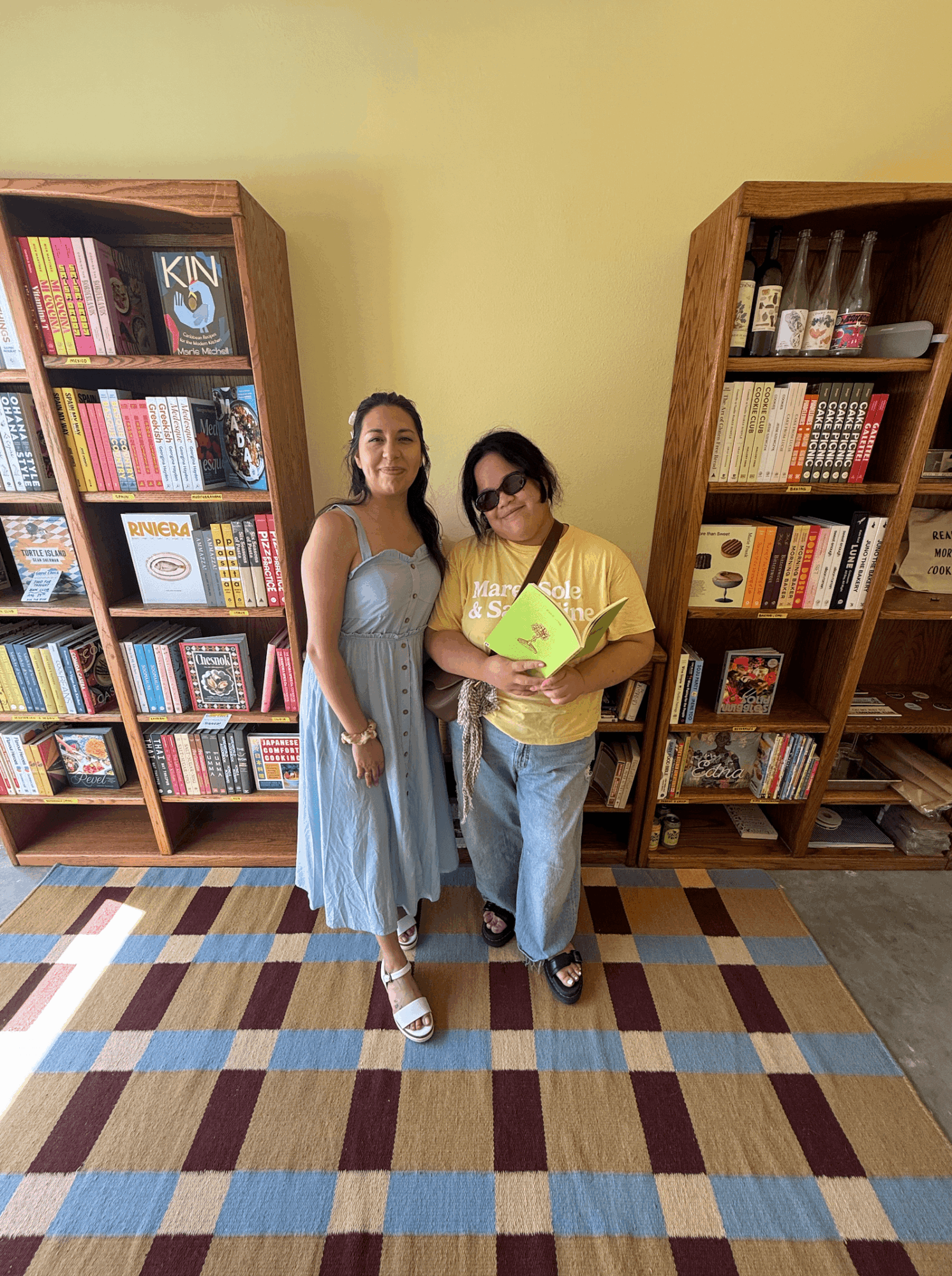 Two women stand in front of bookshelves in a room with a checkered floor, one holding a green book, smiling.