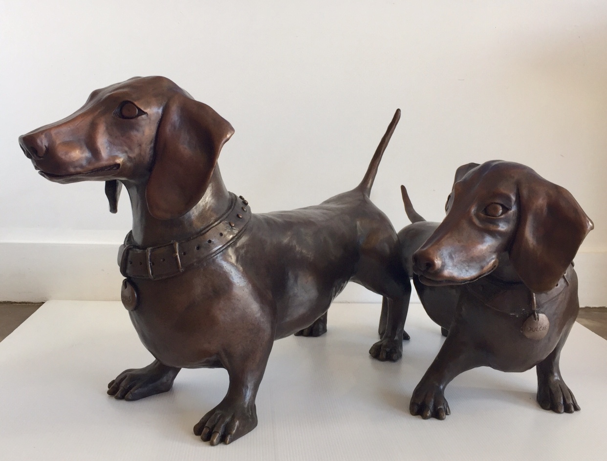 Two realistic dog sculptures, one large and one small, with brown bodies and collars, standing on a white surface.