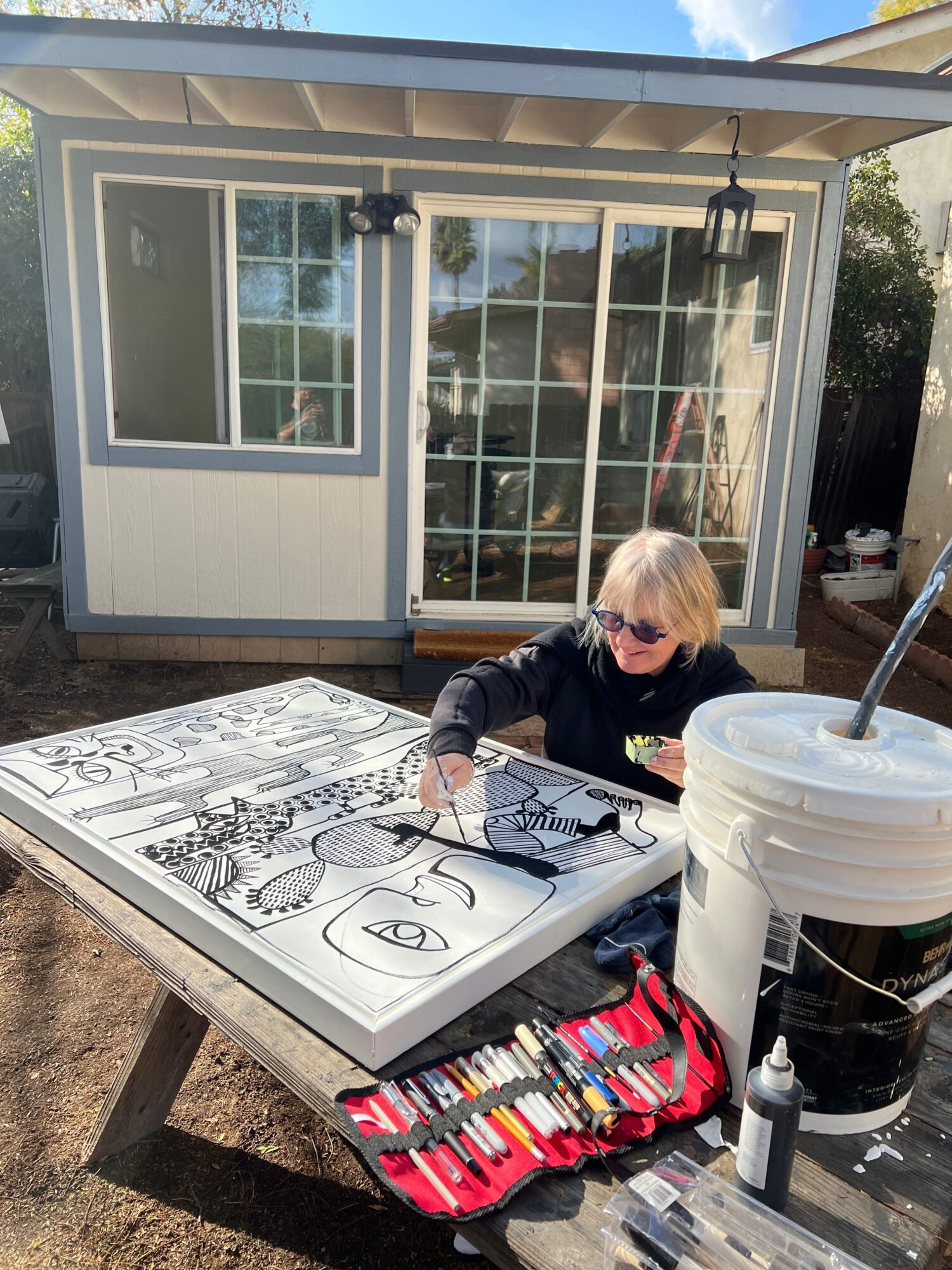 Child coloring on paper at outdoor table with art supplies, backpack nearby, in front of a building with large windows.
