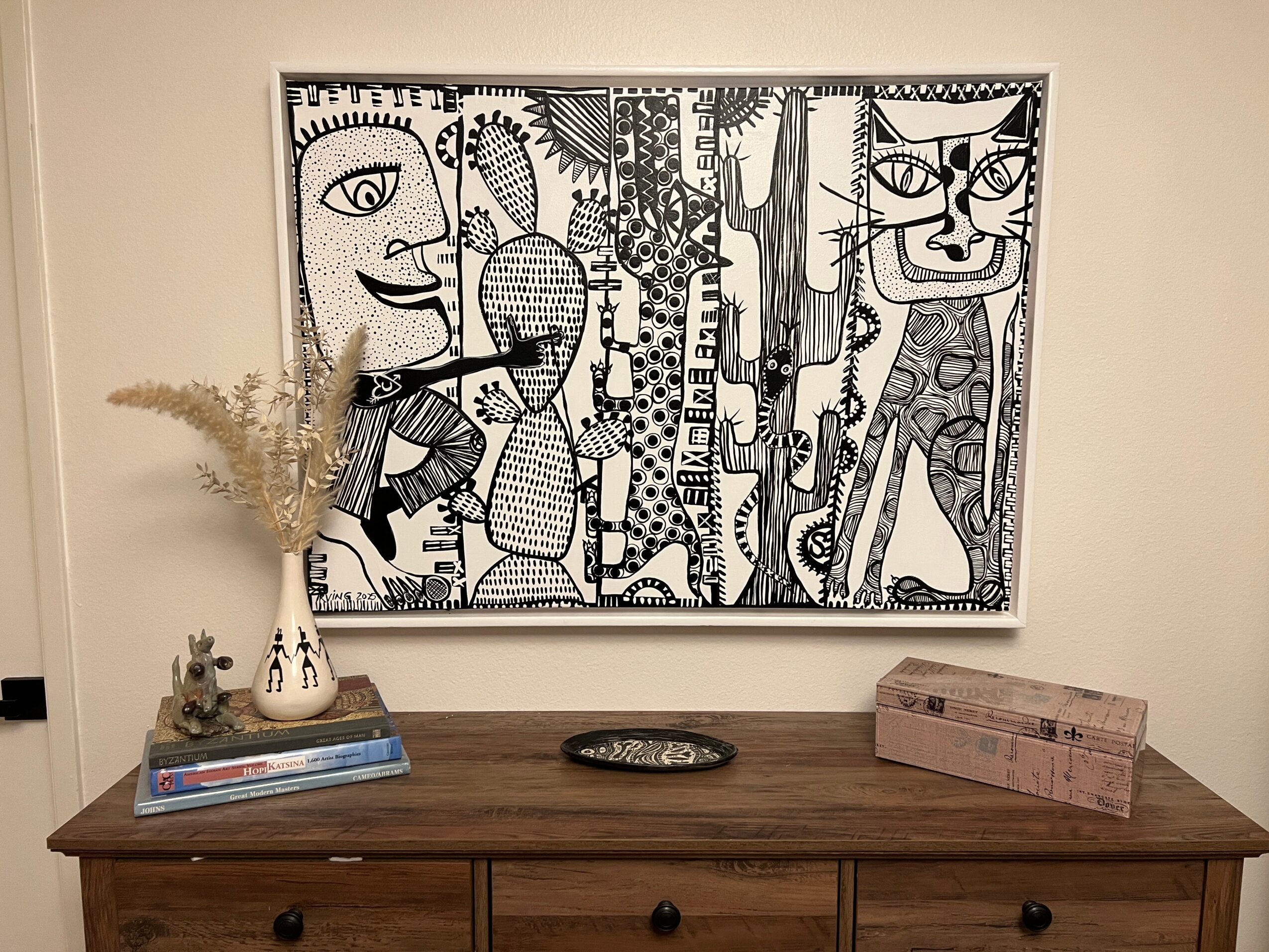 Decorative wall art with abstract faces, a vase with dried plants, books, a decorative tray, and a pink box on a wooden table.