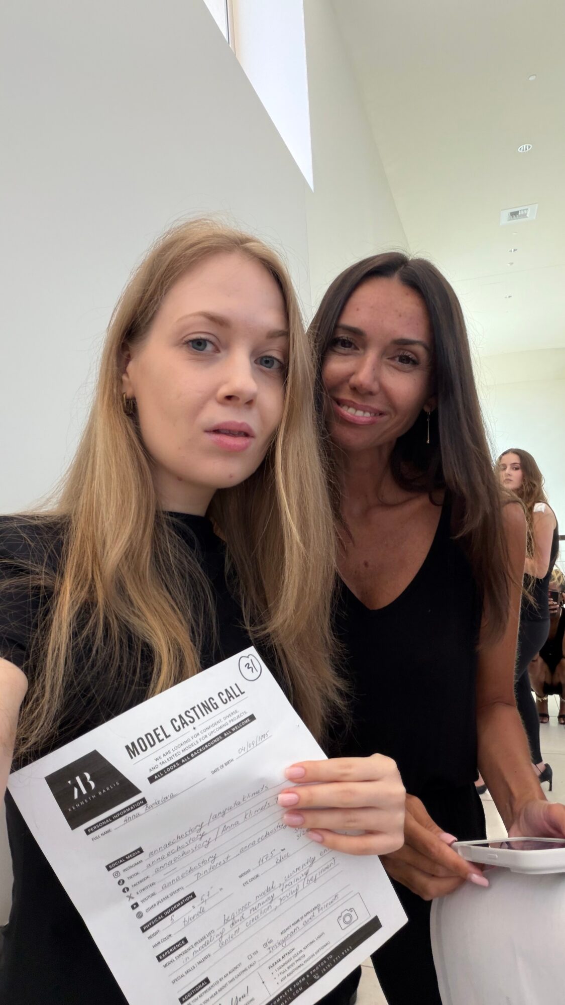 Two women smiling, one holding a document, in an indoor setting with high ceilings and other people in the background.