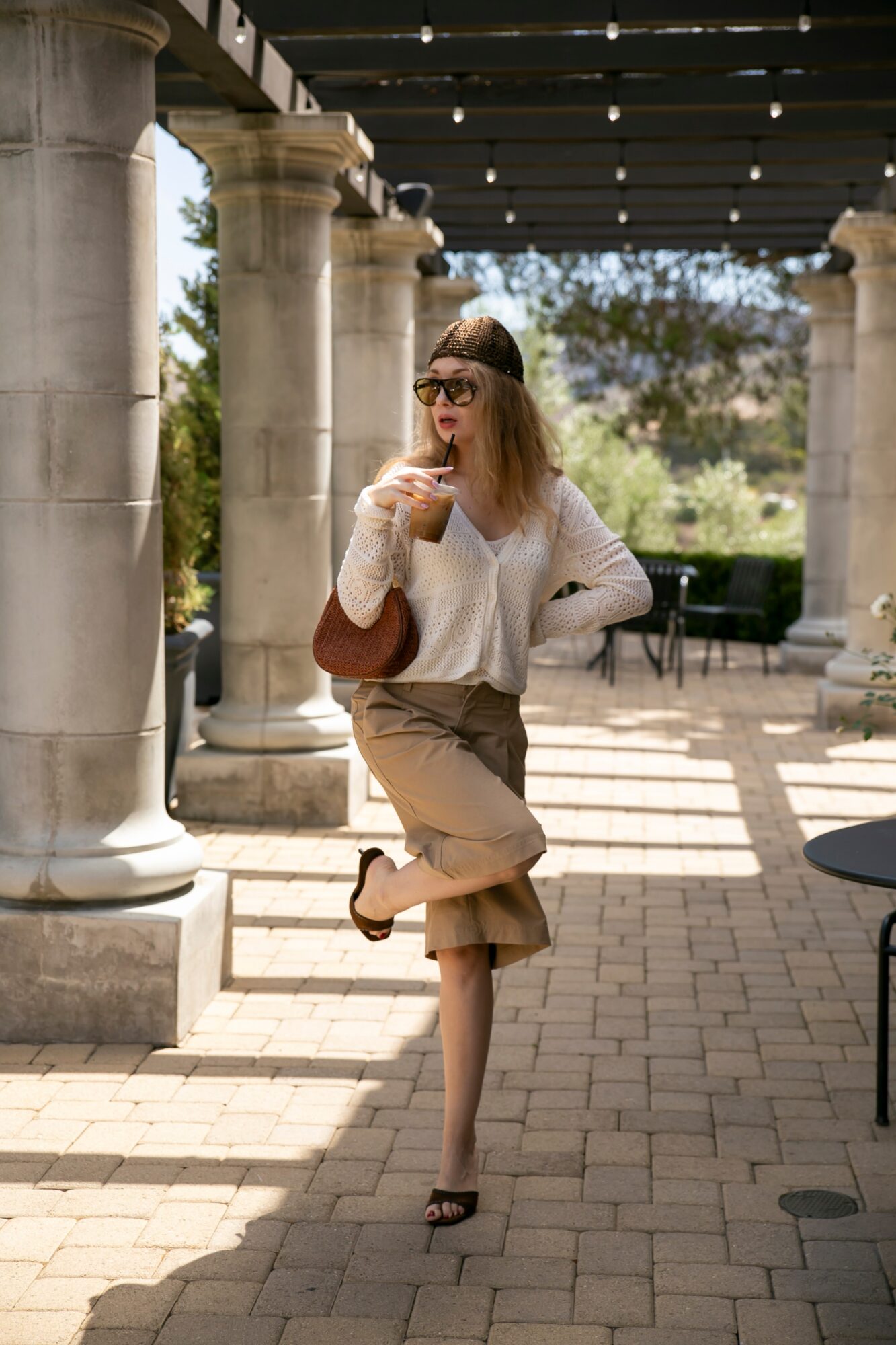 Woman with sunglasses and a white sweater standing outdoors under a pergola, holding a drink, smiling, with trees and chairs in background.