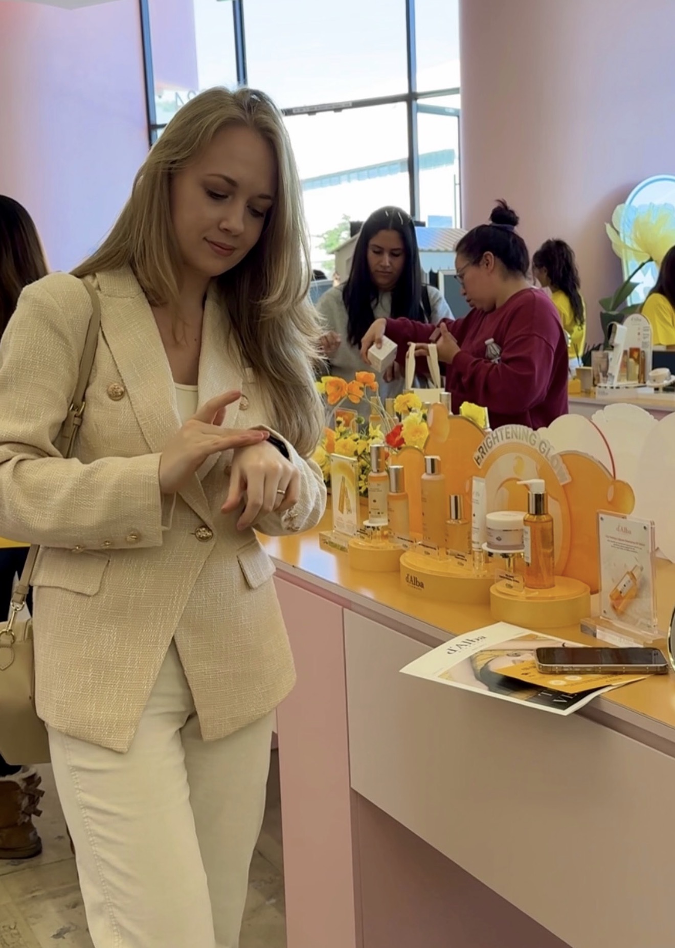 Woman in beige blazer and white pants looking at products on display, with other people in the background, bright window light.