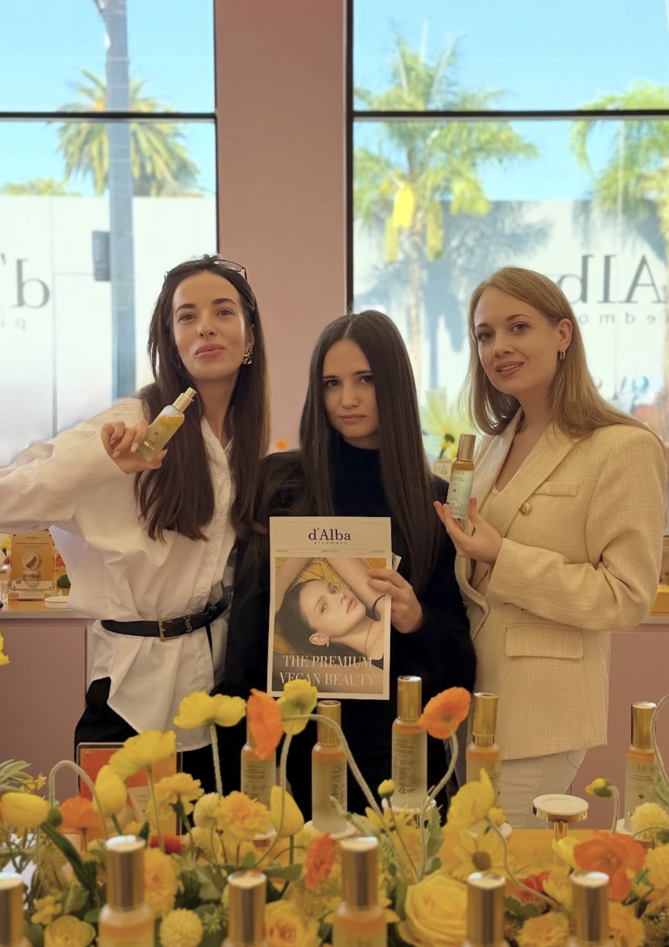 Three women stand behind a table with yellow flowers, holding drinks and a book, in front of large windows with palm trees.