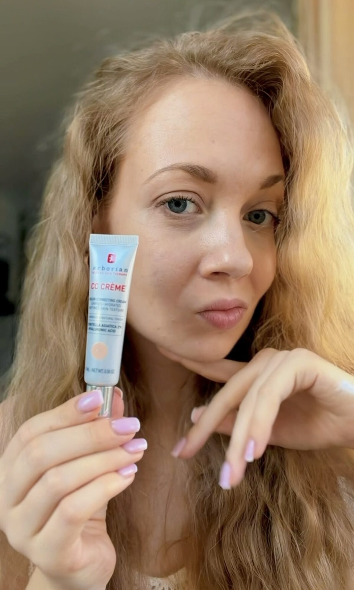 Young woman with long, wavy hair holding a skincare product close to her face, looking at the camera.