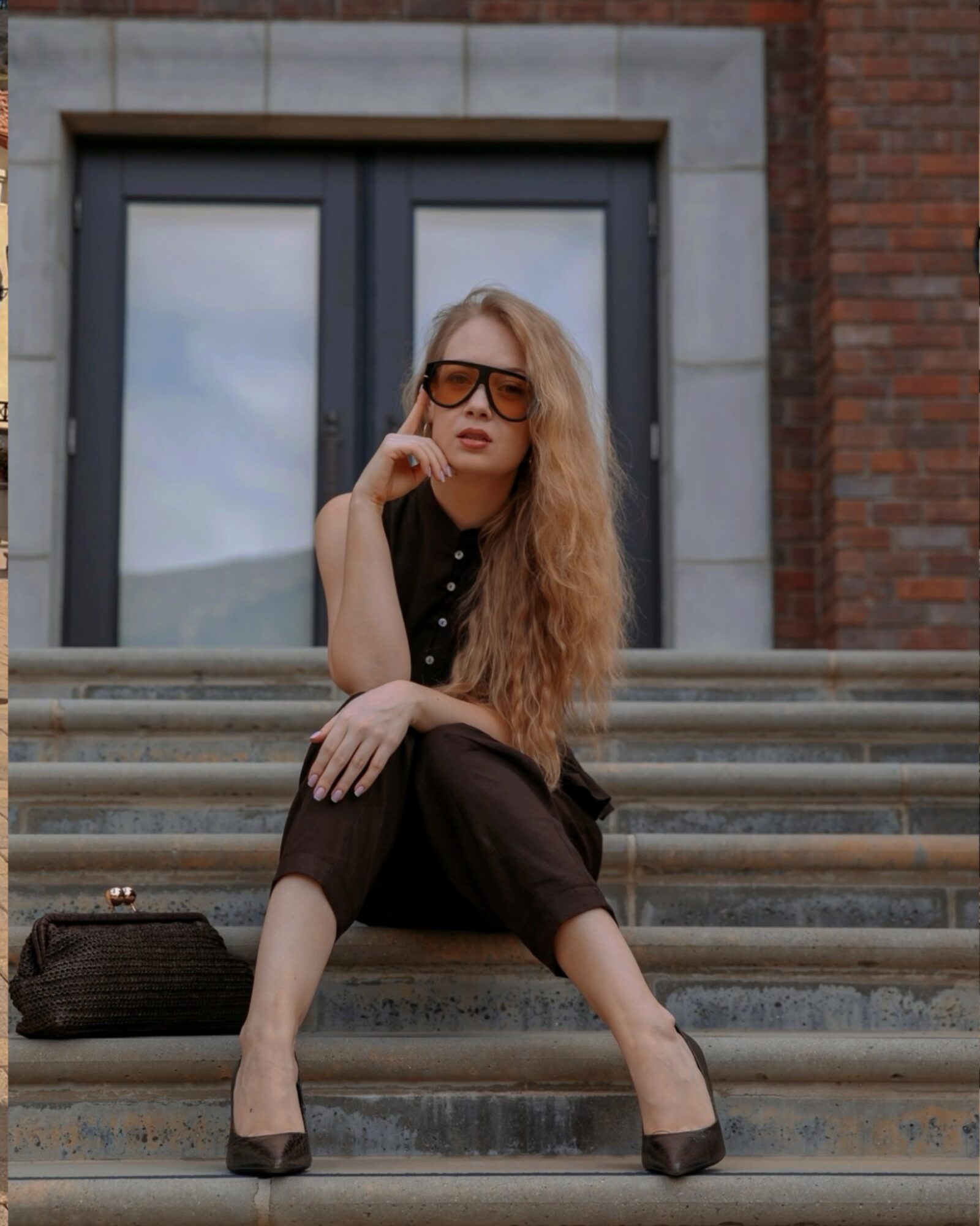 Woman with long hair and sunglasses sitting on stairs outside building, wearing black outfit and heels.