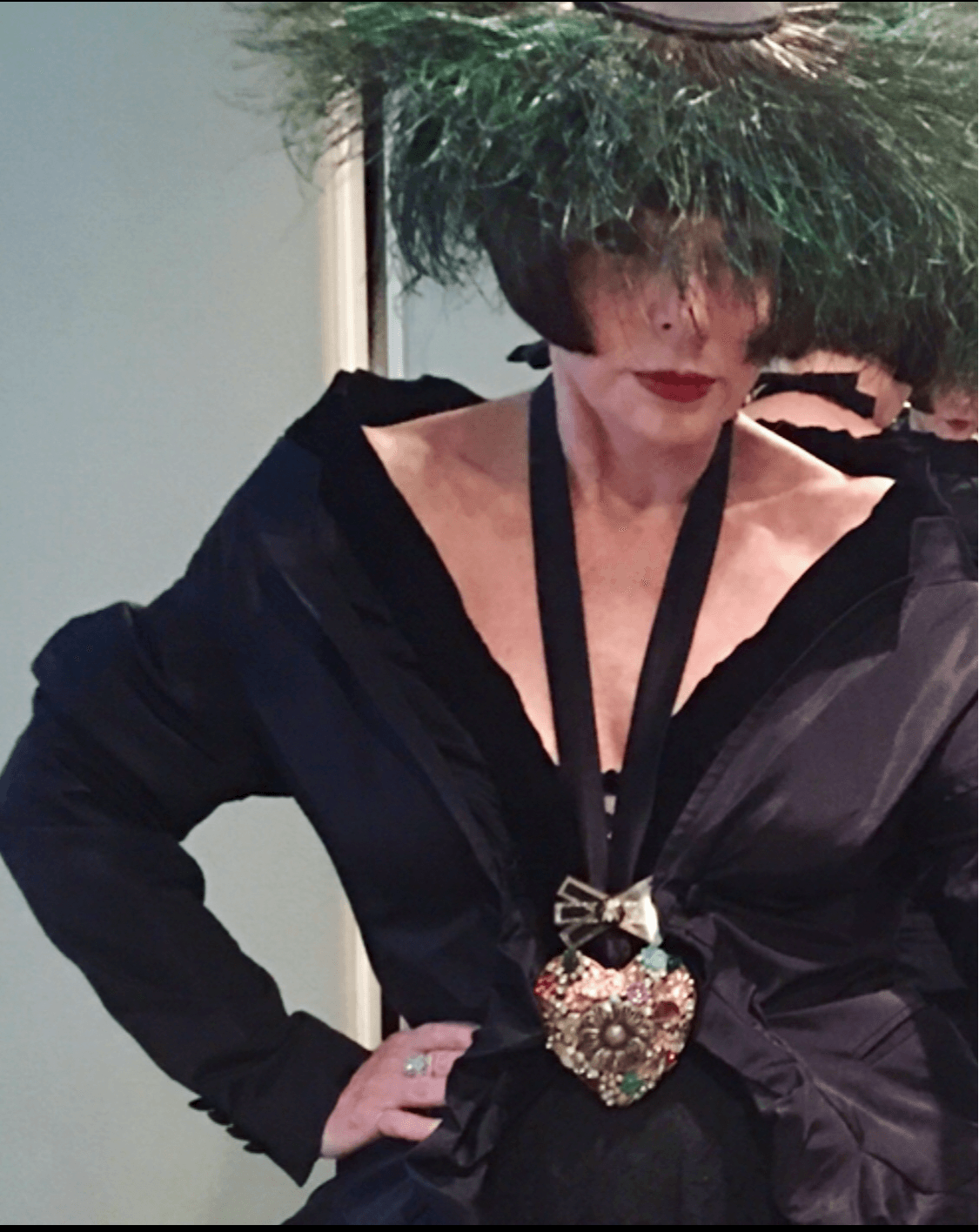 Person wearing a large green hat, black outfit, and a heart-shaped brooch, standing with hand on hip.