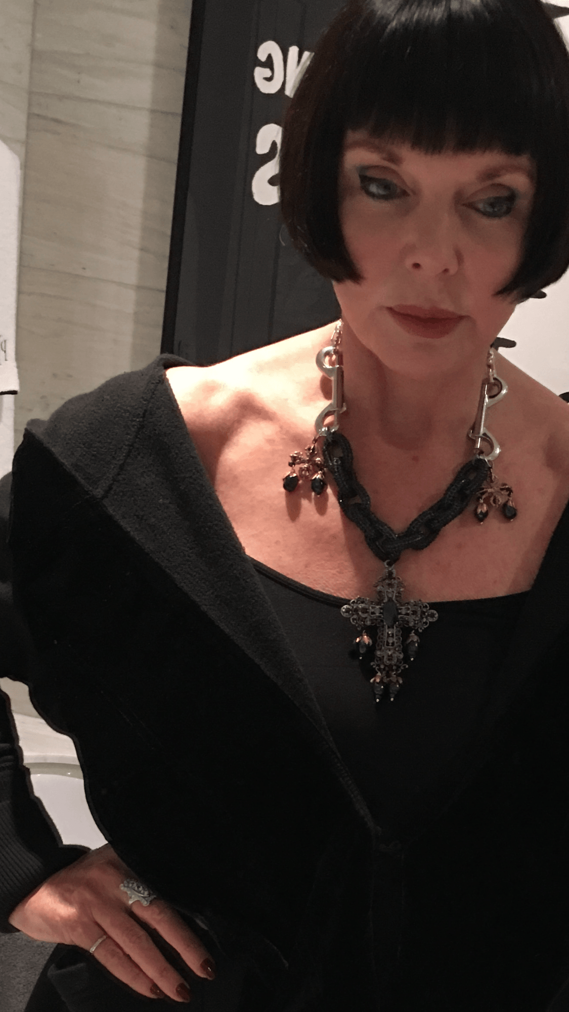 Woman with short black hair wearing a black top and a large necklace, looking down.