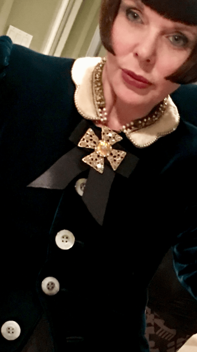 Woman with short dark hair wearing a dark outfit with a pearl and gold brooch, and a pearl necklace.