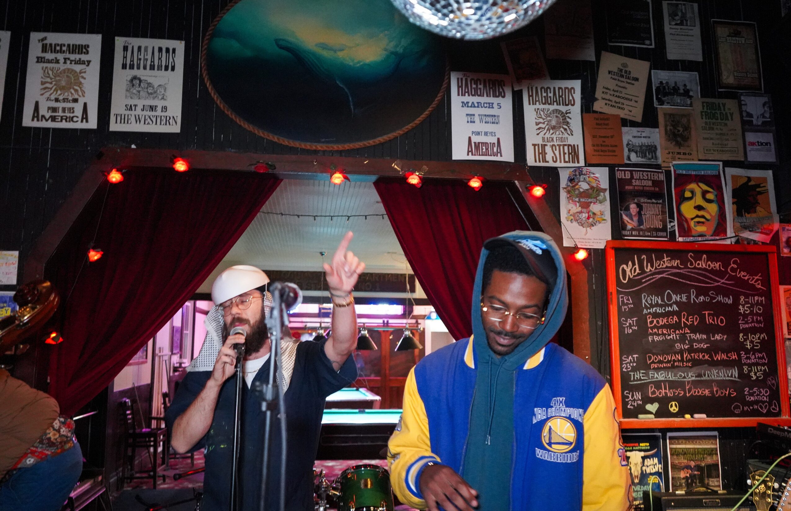 Two men perform in a bar with red curtains, posters, and a chalkboard menu behind them. One sings into a microphone, the other looks down.