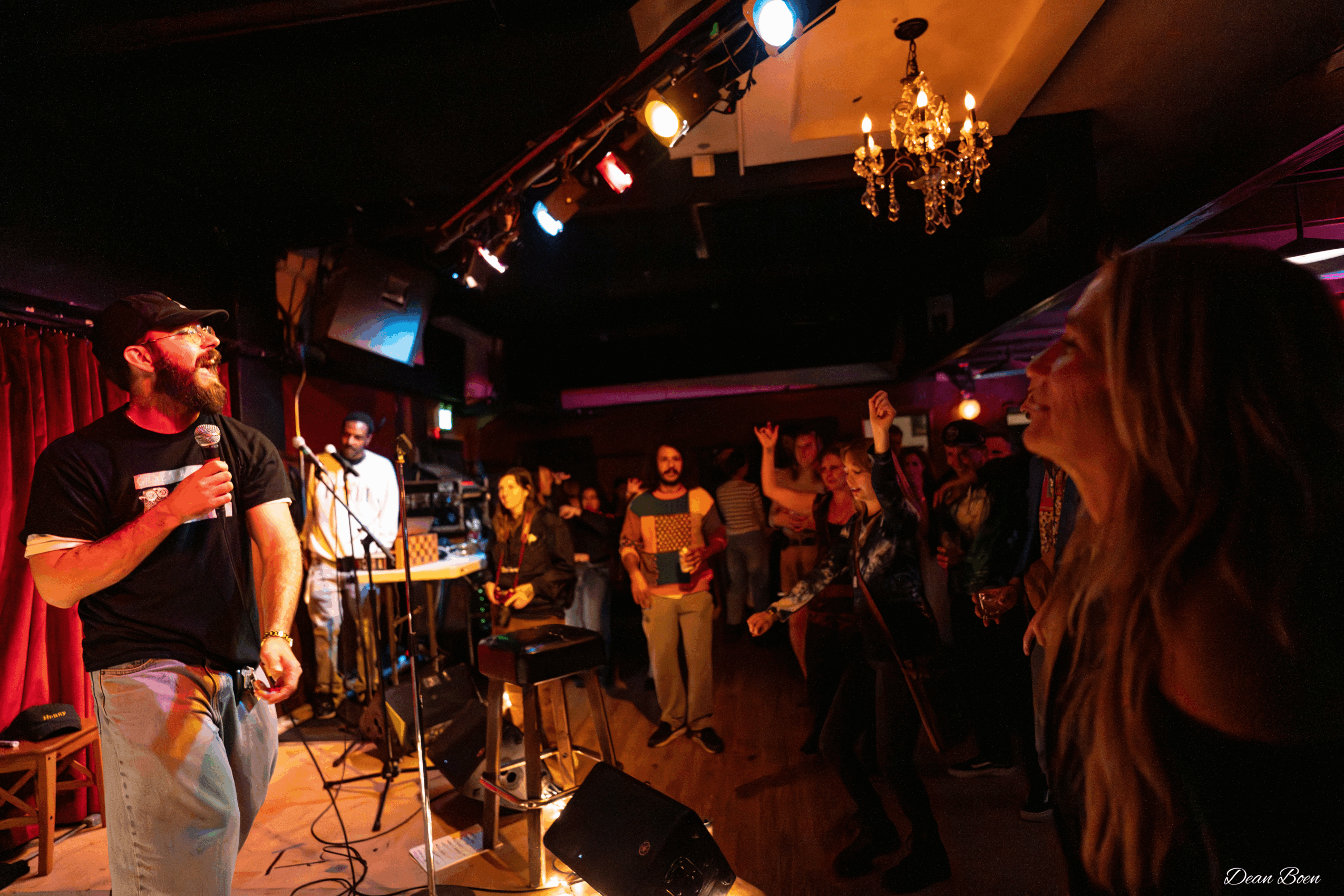 Person singing into a microphone on stage with a band, audience watching in a dimly lit venue with colorful lights.