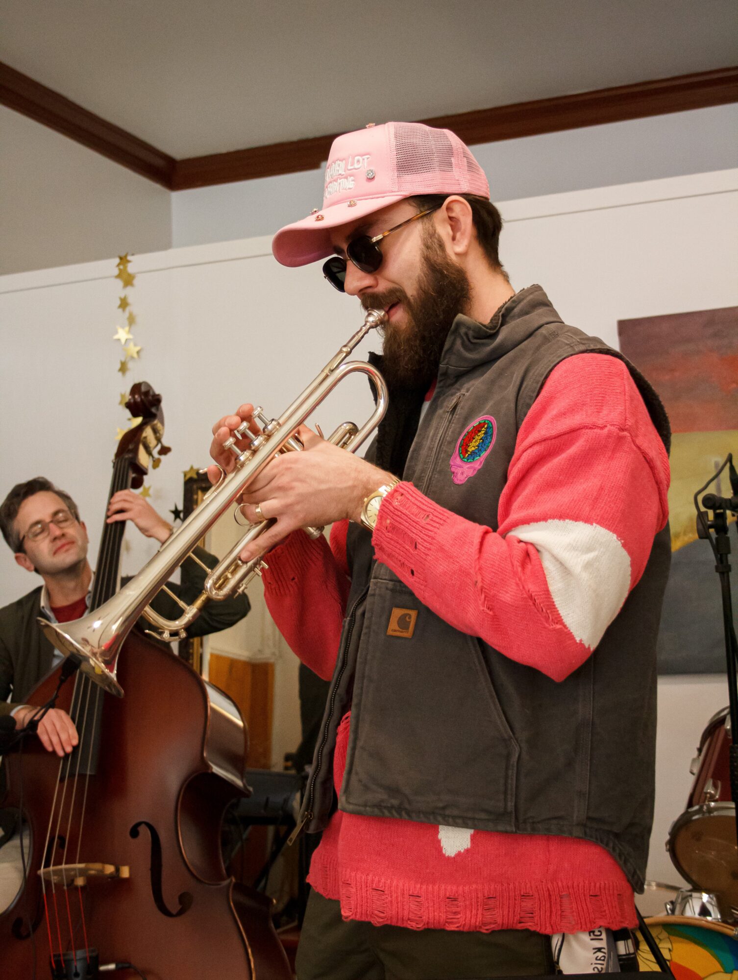 Man playing trumpet, wearing pink cap, sunglasses, and a colorful jacket, with a double bass player in the background.