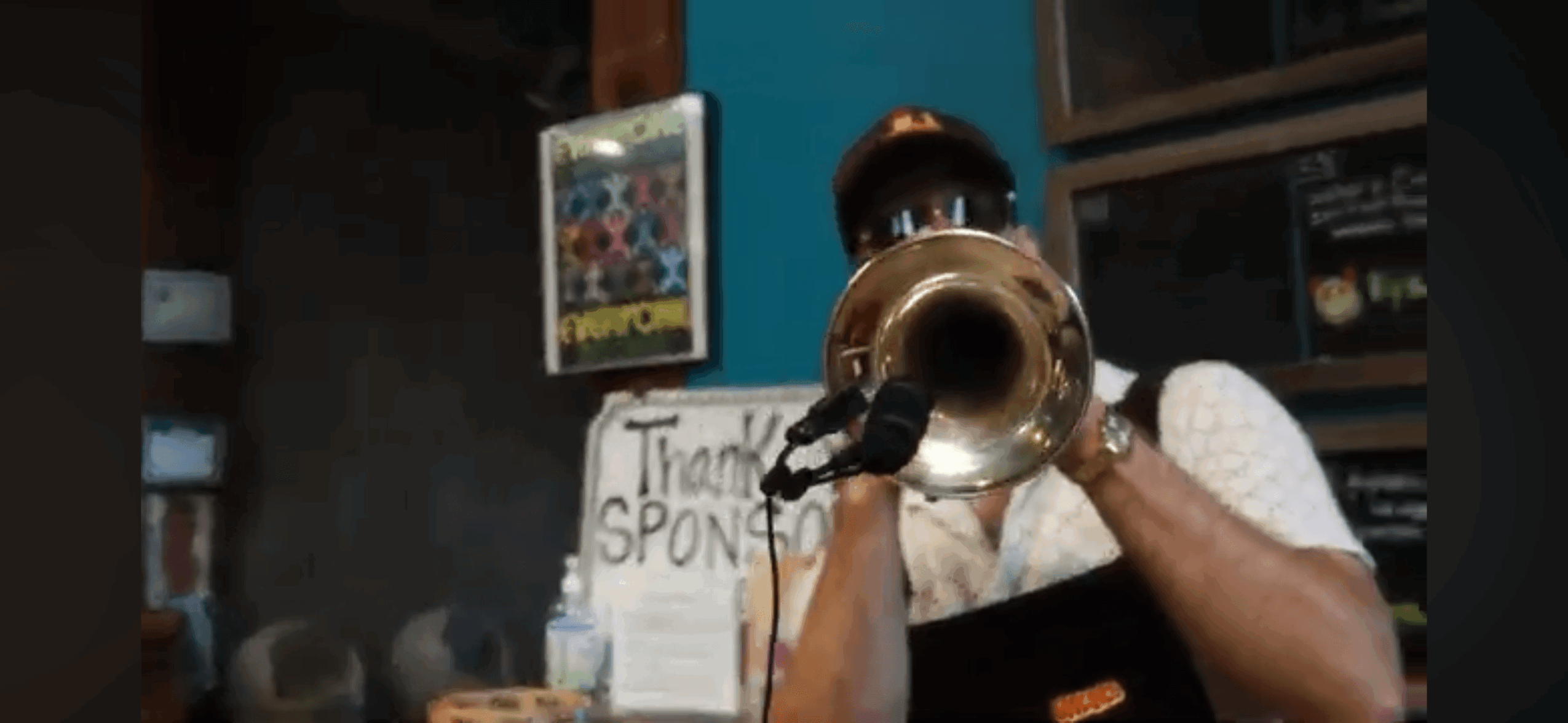 Person playing a brass instrument in a room with a sign and shelves, partially obscured face, wearing a white shirt.