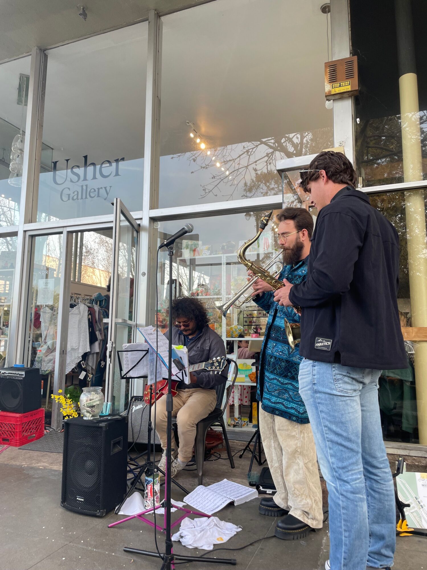 Three people stand outside a glass storefront, with one person playing a guitar and others watching, papers on the ground.