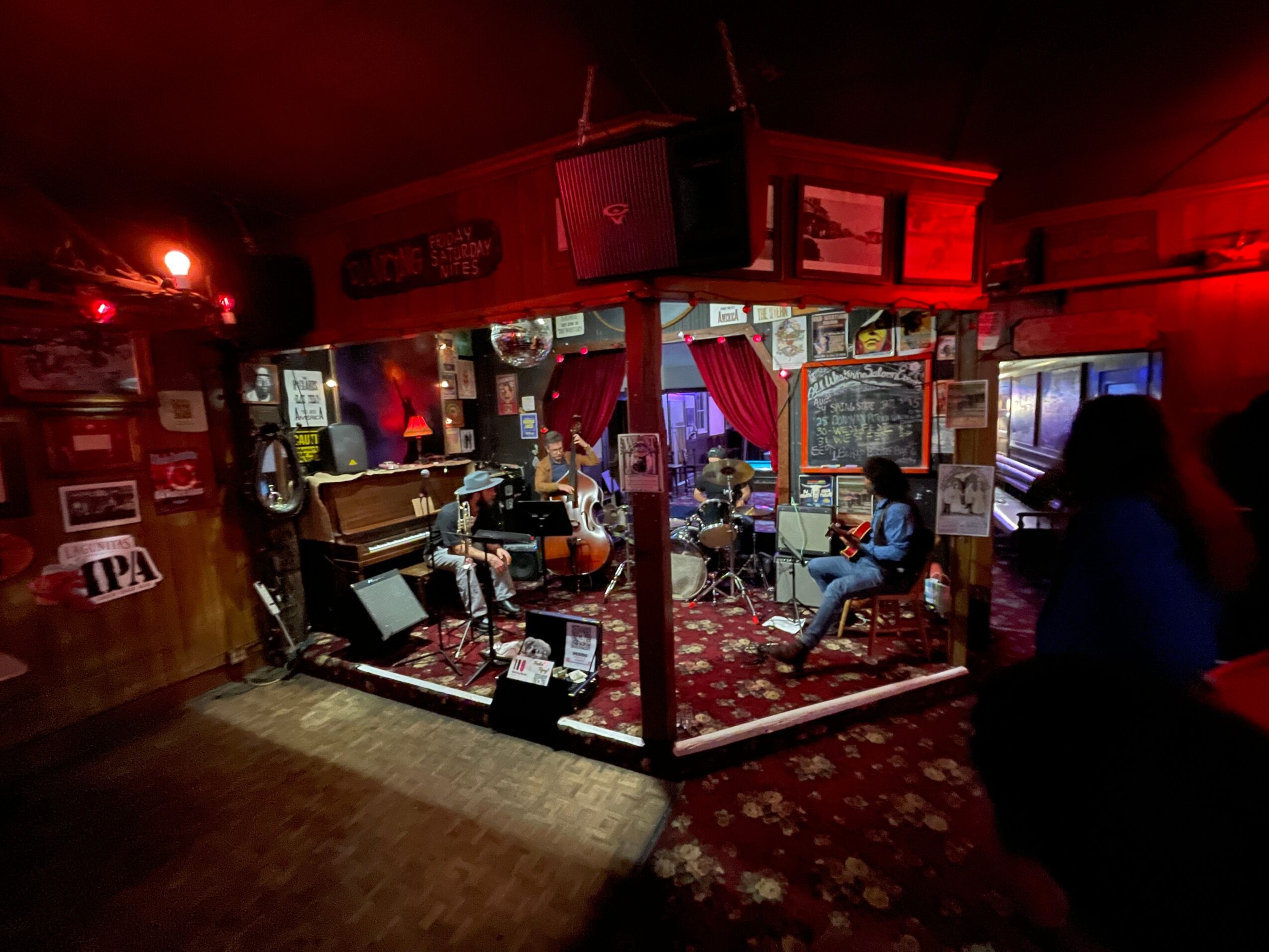 Music venue with people sitting and playing instruments, decorated with posters and red curtains, dim lighting, and a patterned carpet.