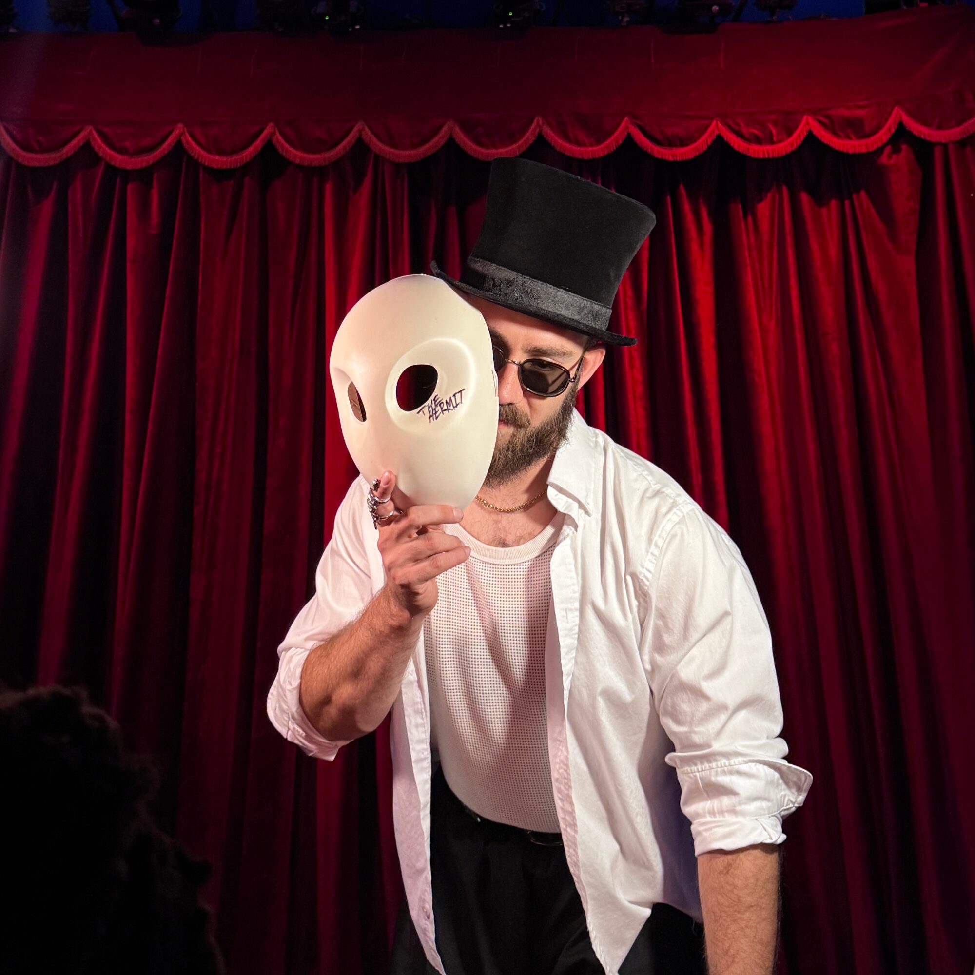 Person wearing a top hat and sunglasses holding a white mask in front of red curtain backdrop.
