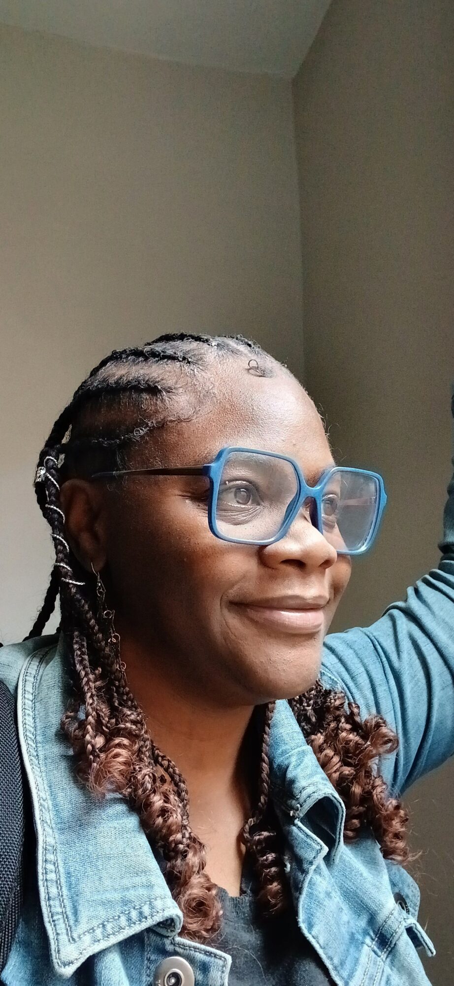 Person with glasses and braided hair looking upward, wearing a denim jacket, against a plain background.