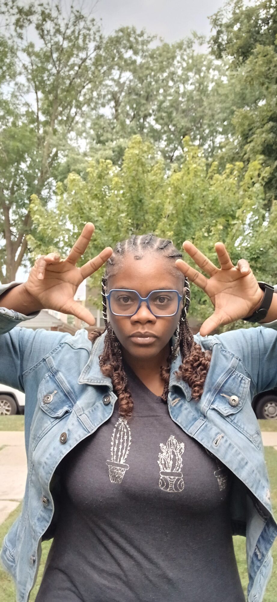 Person outdoors making peace signs with both hands, smiling, with trees in the background.
