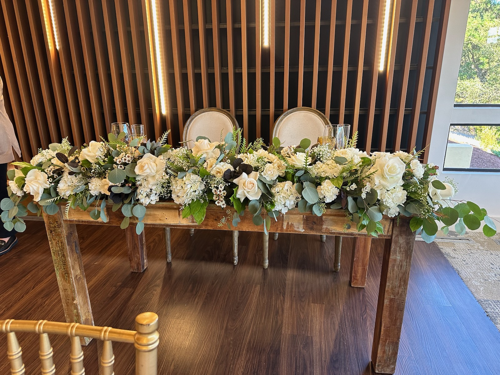 Long wooden table decorated with white flowers and greenery, two chairs behind, wooden slat wall and window in background.