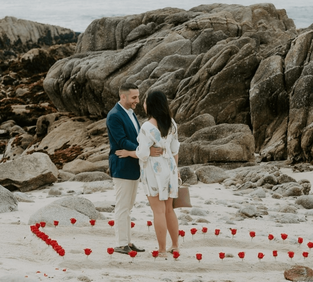 A couple stands on a sandy beach surrounded by rocks, holding hands, with red rose petals arranged in a heart shape around them.