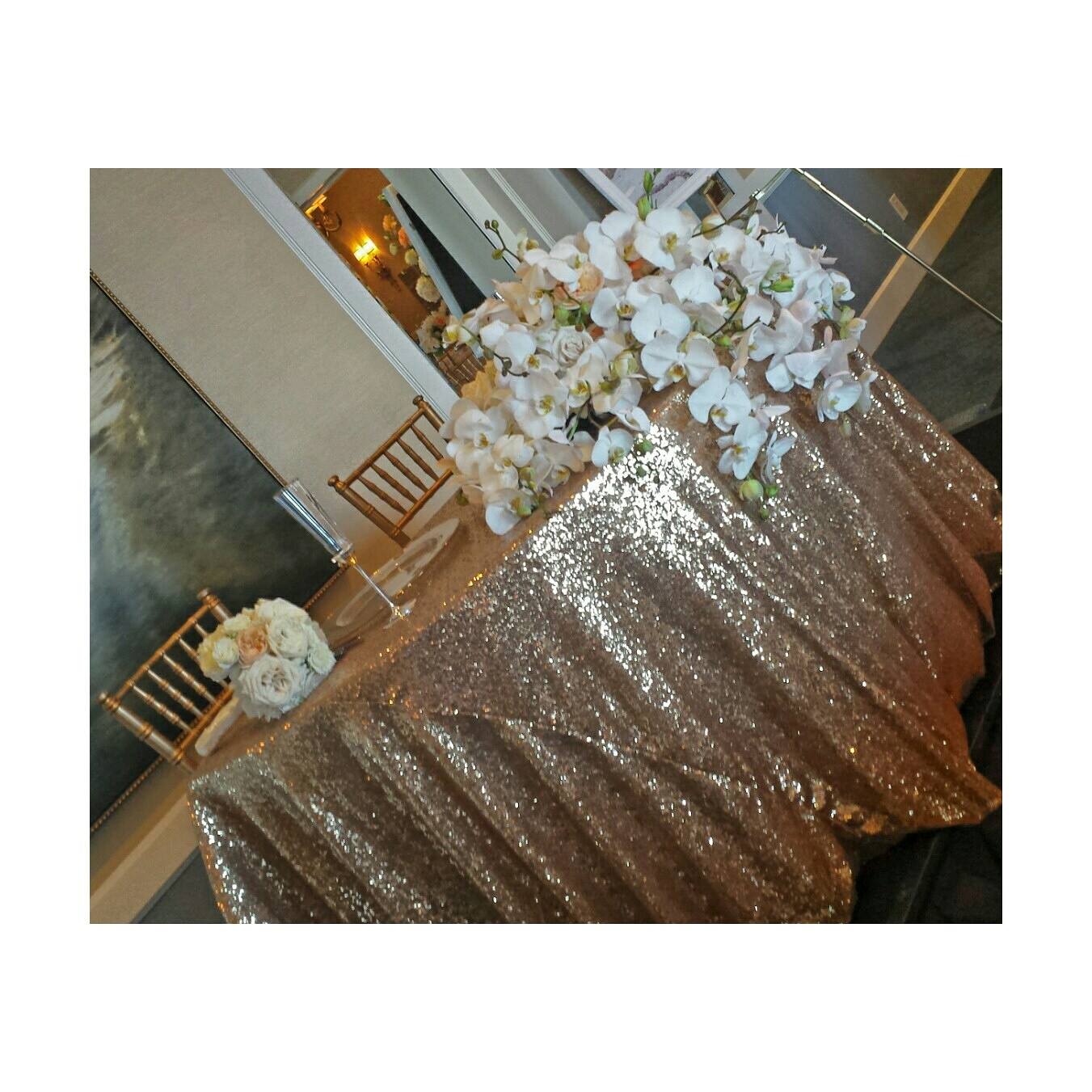 Table with a glittery gold tablecloth and a large floral centerpiece of white flowers, set in a decorated room.