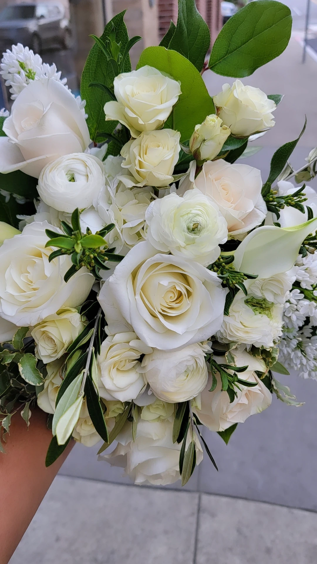 Bouquet of white roses, lilies, and small white flowers with green leaves, held outdoors.